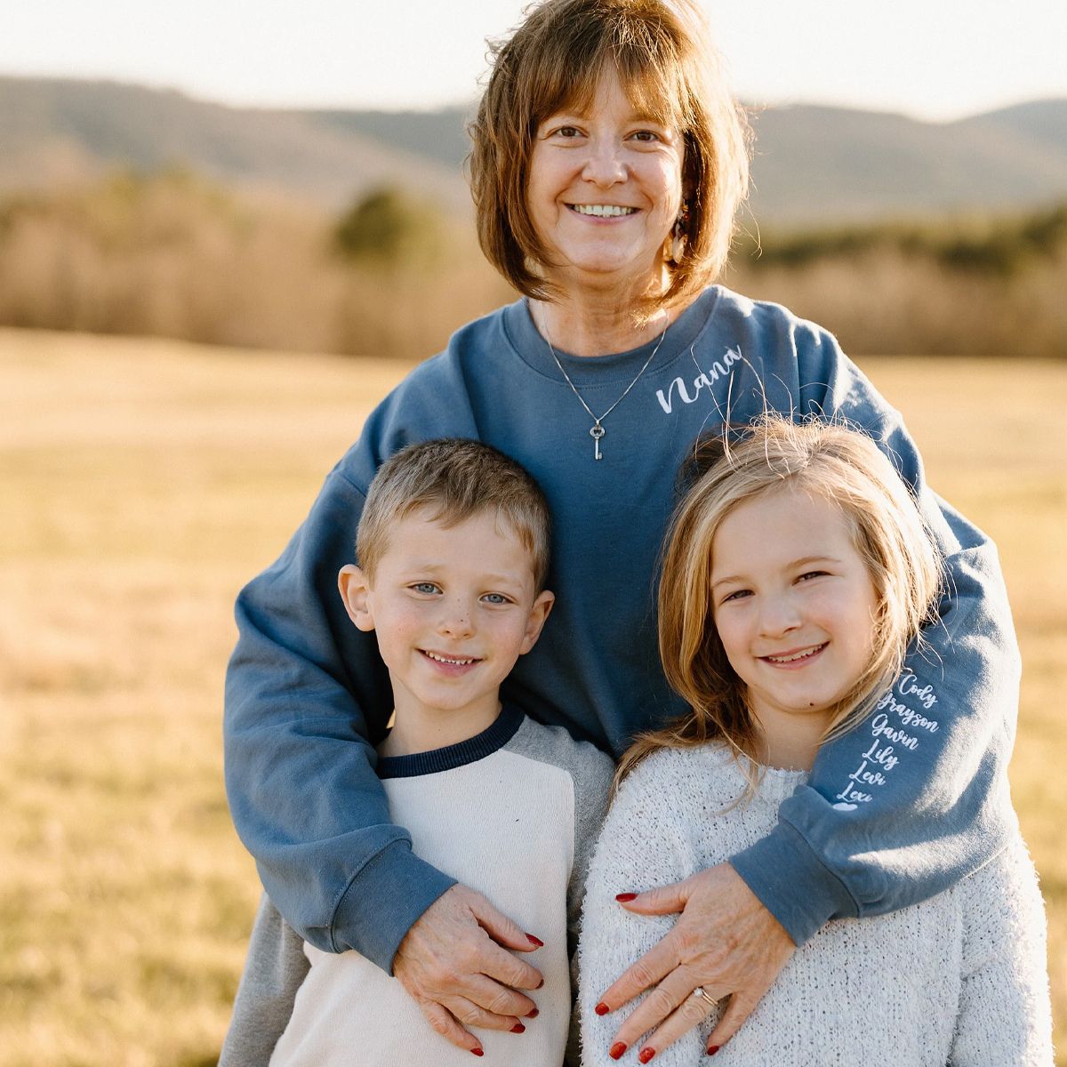 Smiling grandmother wearing a blue 'Nana' sweatshirt with white embroidered text, embracing two young grandchildren in a scenic outdoor field.