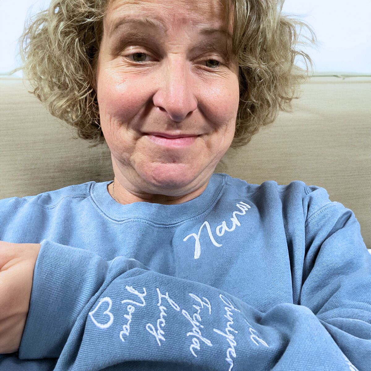 Smiling grandmother with curly blonde hair in a light blue 'Nana' sweatshirt, showing embroidered grandkids' names on the sleeve.