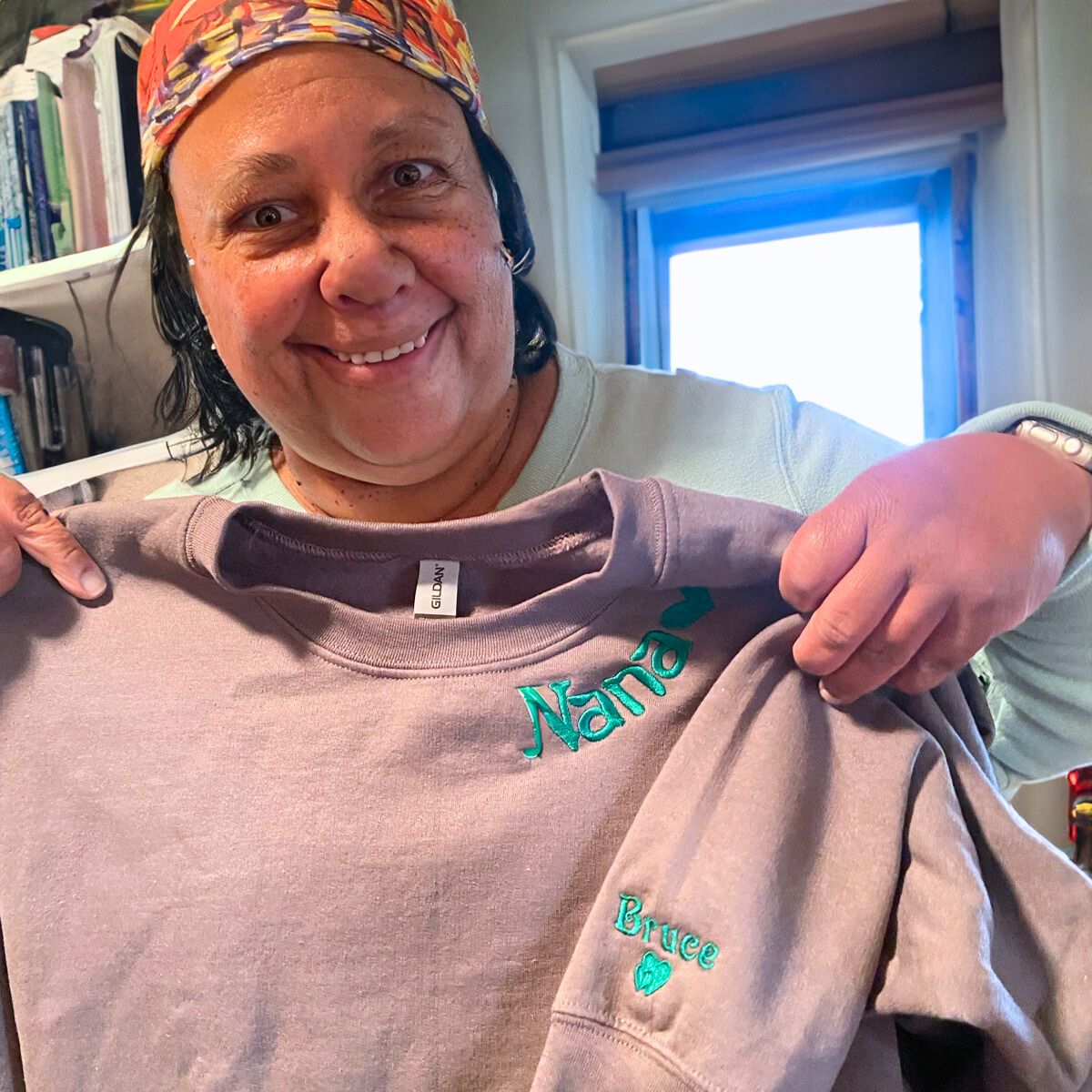 Grandmother in a mauve 'Nana' sweatshirt with green embroidered text and a grandchild's name, holding it up with a bright smile.