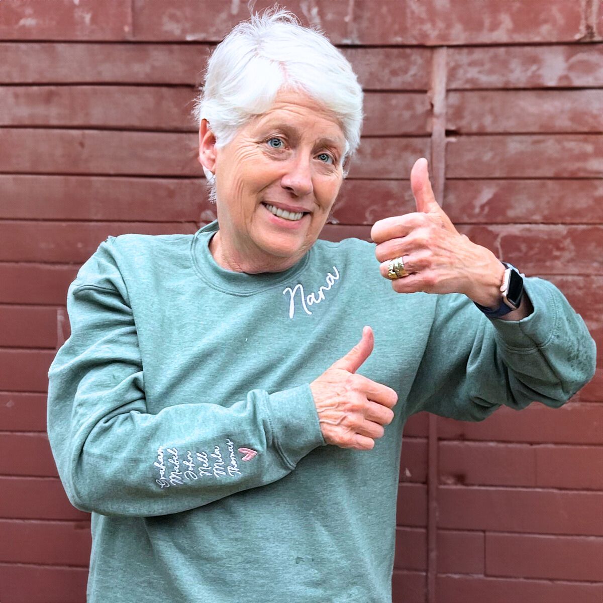 Smiling grandmother in a sage green 'Nana' sweatshirt, giving a thumbs-up pose against a rustic red wooden fence.