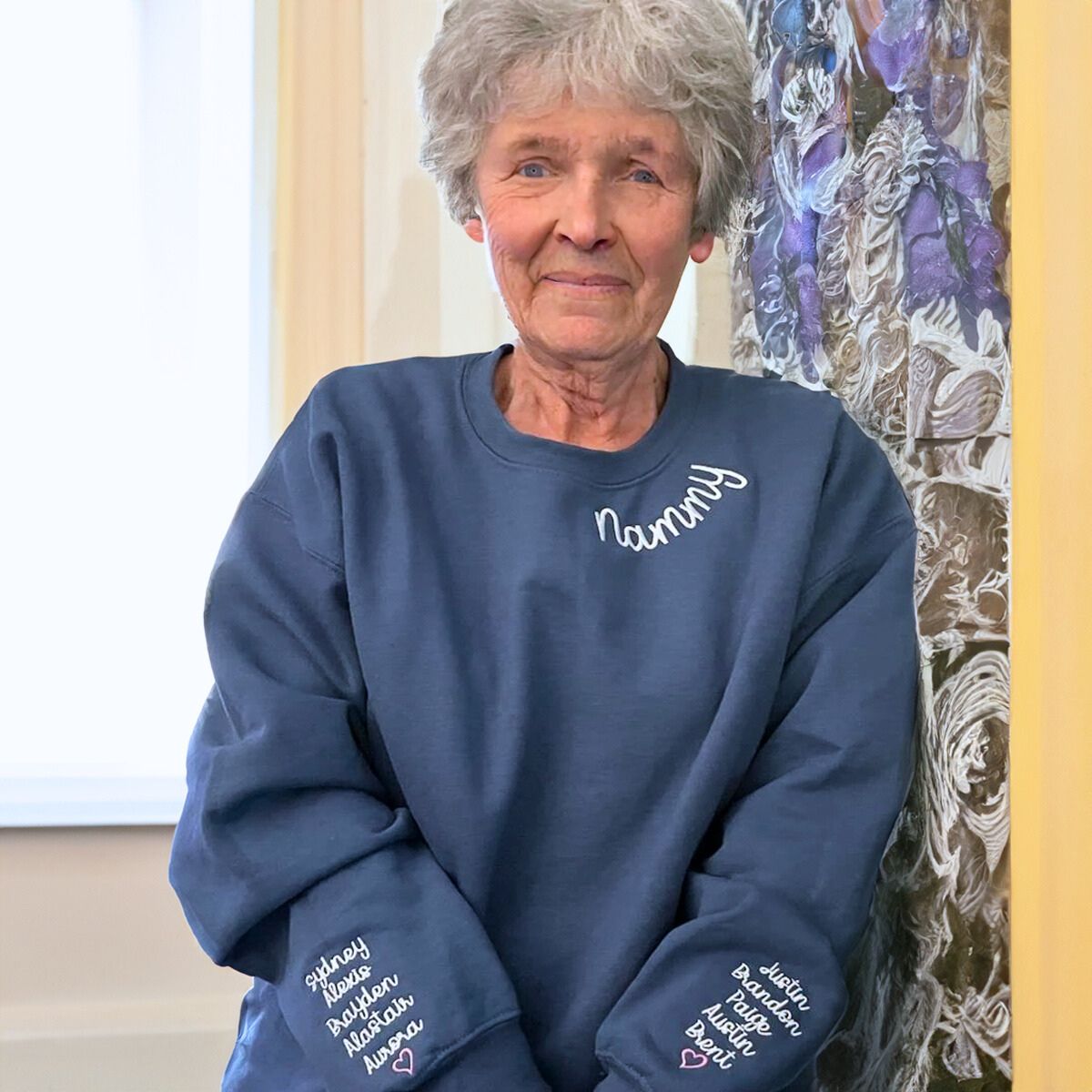 Elderly woman with short gray hair wearing a navy blue 'Nanny' sweatshirt with white embroidered text and personalized grandkids' names on the sleeves, posing indoors.