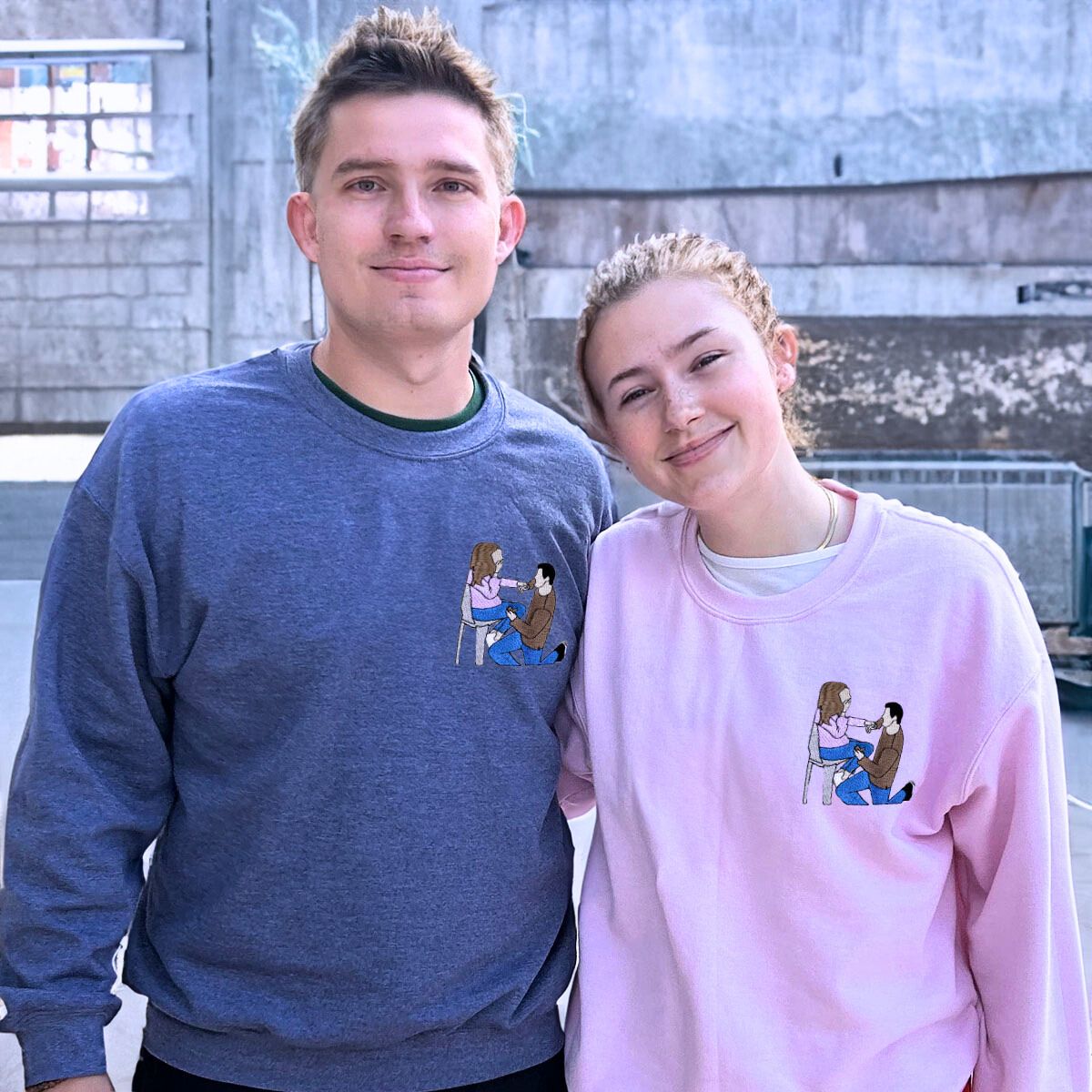 Playful couple wearing matching blue and pink Embroly sweatshirts with custom embroidered couple design.