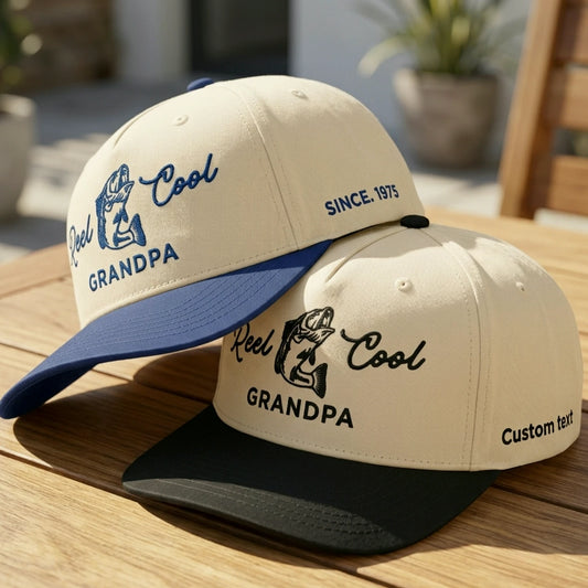 Two baseball caps displayed on a wooden table in a sunlit outdoor setting. The front cap is cream with a dark blue brim and blue embroidered text reading 'Reel Cool GRANDPA' and 'SINCE 1975.