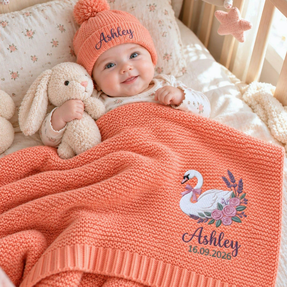 Smiling baby with a personalized coral knit blanket featuring swan and floral embroidery, a birth date, and a matching beanie, an idea for a newborn gift.