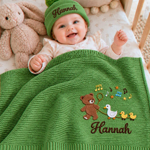 Smiling baby under a personalized green knit blanket embroidered with a teddy bear, ducks and musical notes, wearing a matching beanie, a baby shower gift.