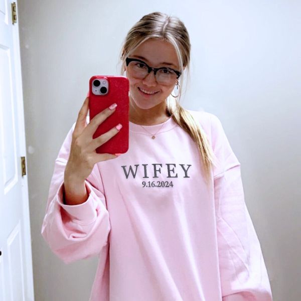 Smiling woman in light pink 'Wifey' sweatshirt with custom date, taking a mirror selfie.