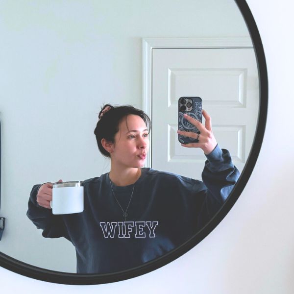 Woman in navy blue 'Wifey' sweatshirt taking a mirror selfie while holding a coffee mug.