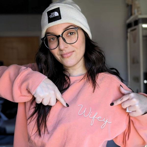 Woman in pink 'Wifey' sweatshirt and beanie, pointing at the embroidered text.