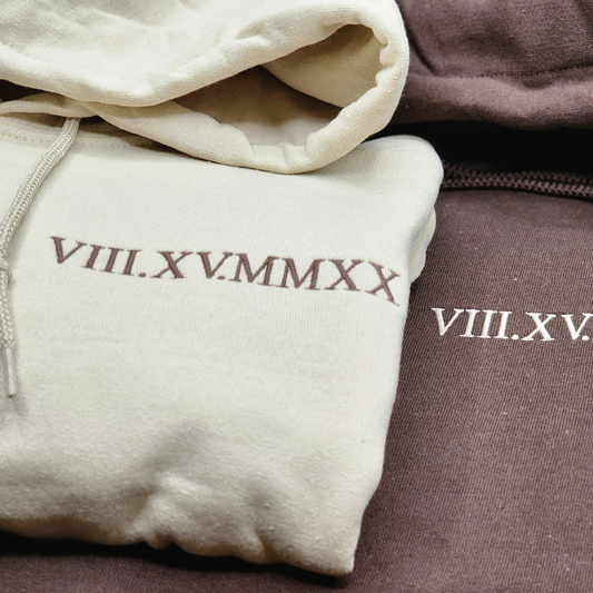 Cream drawstring hoodie folded beside a mauve hoodie, both with Roman numeral wedding date embroidered on the pocket